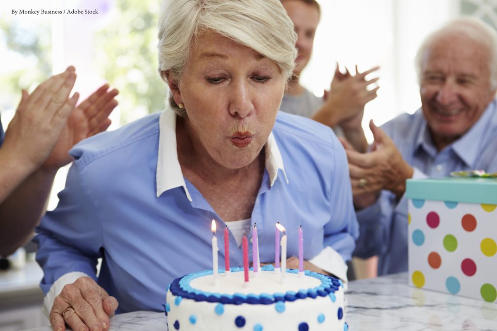 Senior Woman Blows Out Birthday Cake Candles At Family Party